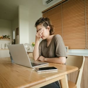 A woman is utilizing EFT to tap on specific points on her face for headache relief while sitting at a table with a laptop.