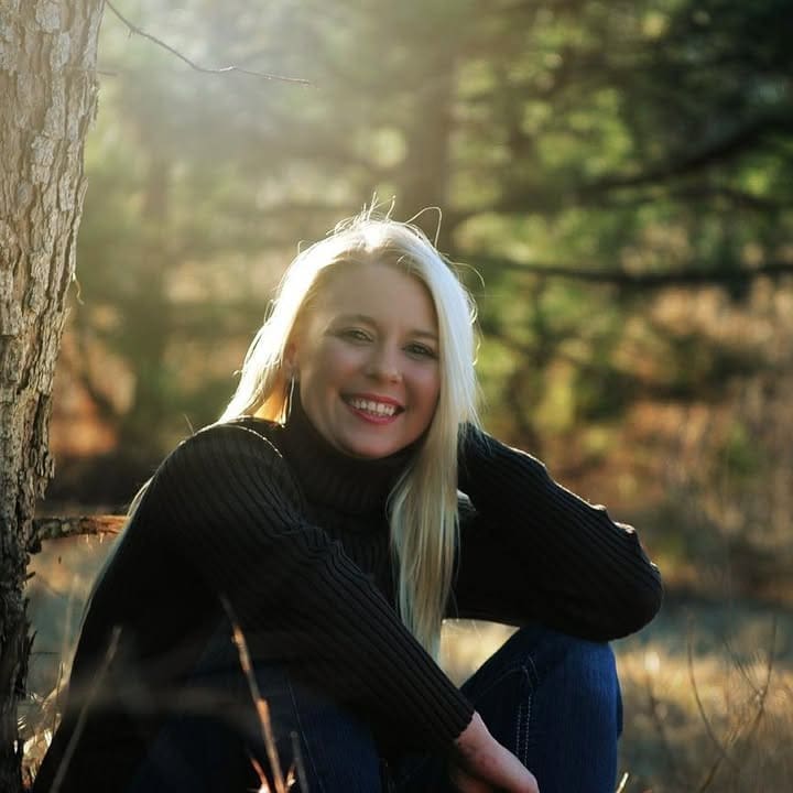 A person with long blonde hair wearing a black sweater sits on the ground by a tree, smiling and practicing tapping, with sunlight and trees in the background.