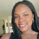 A woman with braided hair smiles at the camera indoors, wearing a sleeveless top and a necklace.
