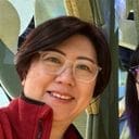 A person with short dark hair, glasses, and a red jacket smiles at the camera. Green metallic shapes are visible in the background.