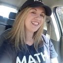 A person with long blonde hair smiles in the driver's seat of a car, wearing a dark hat and a dark shirt with the word "MATH" printed on it.
