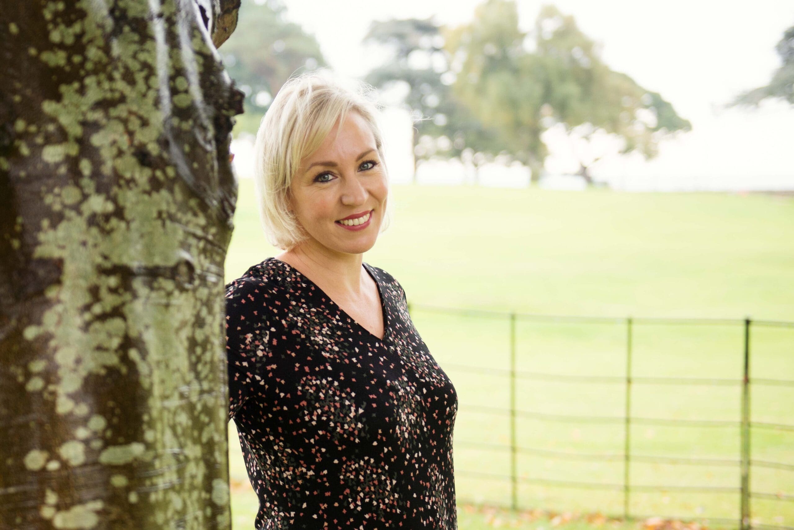 A woman with short blonde hair stands next to a tree, smiling, in a park with a grassy field and trees in the background.