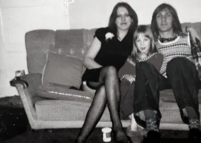 A woman, a man, and a child sit closely together on a sofa in a living room. The photo is black and white and appears to be vintage.