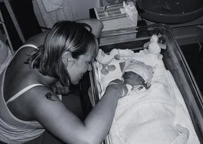 A woman smiles and leans over a hospital crib, touching the hand of a newborn baby wrapped in a blanket, with stuffed animals nearby.
