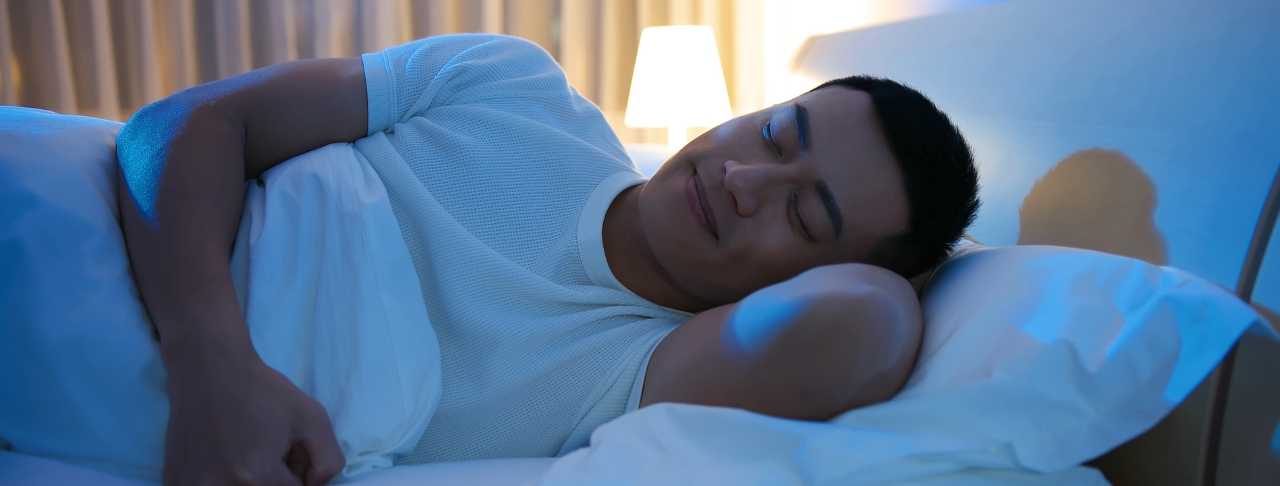 A person in a white shirt is lying in bed, smiling with eyes closed, appearing to be asleep. A lit lamp and curtains are visible in the background.