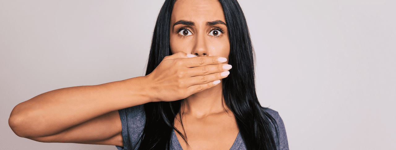 A woman with long dark hair covers her mouth with her hand, looking directly at the camera with wide eyes.