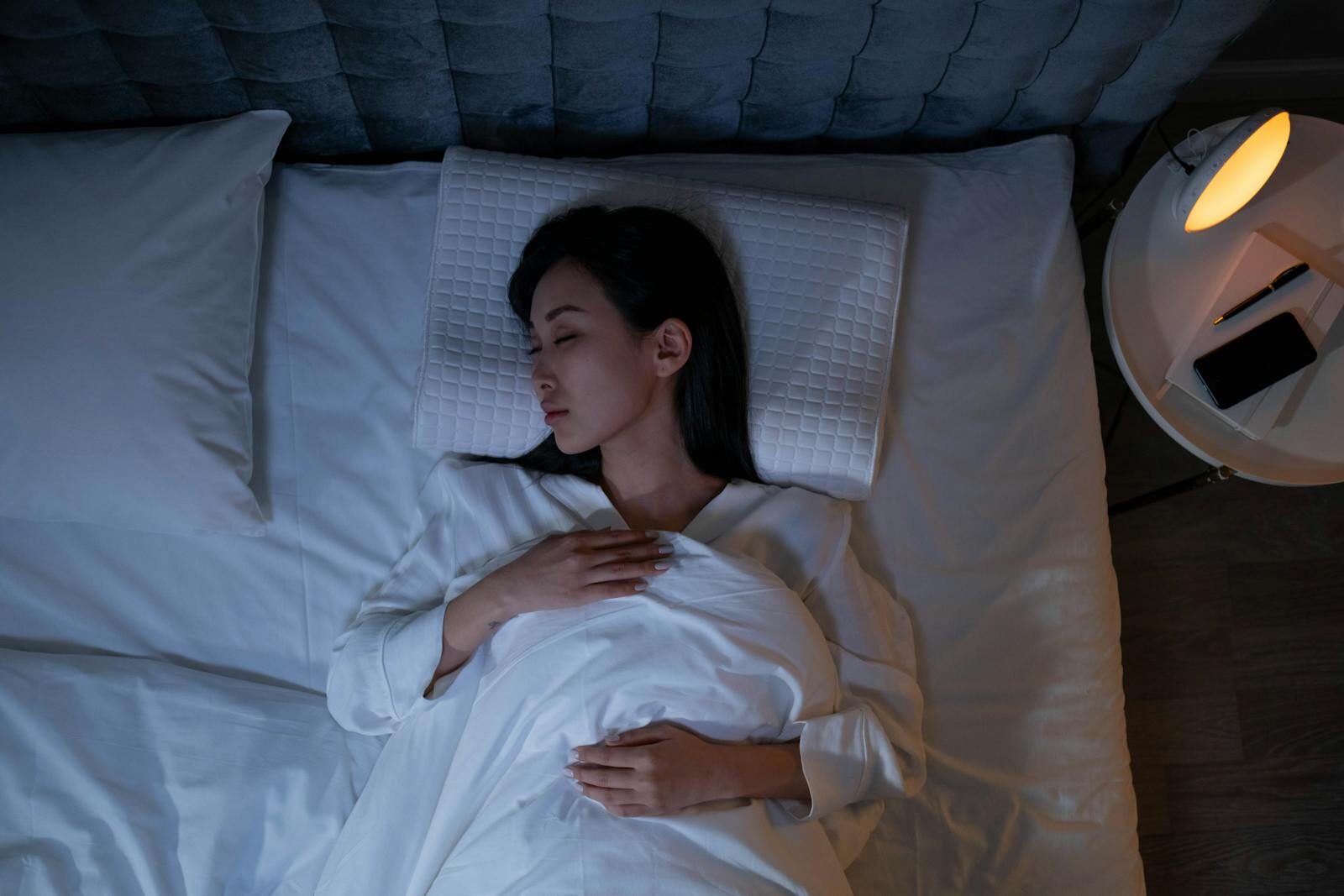 Woman lying on her back in bed with eyes closed, practicing EFT tapping for sleep to calm her racing mind, covered with a white blanket, one hand on her chest, next to a nightstand with a lamp, phone, and glasses.