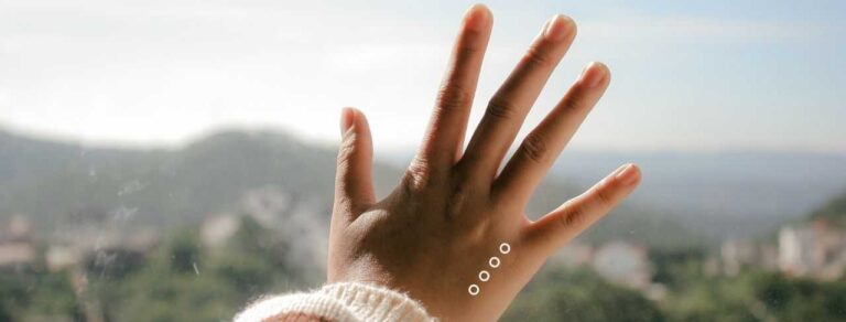 A hand with a white sweater sleeve is held up against a window, with four small white circles on the skin and a blurry landscape in the background.