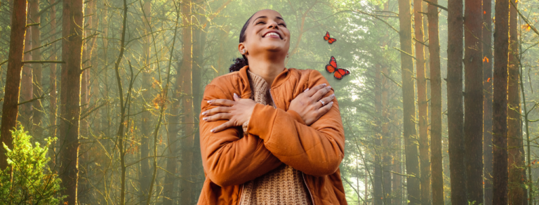 A person in an orange jacket stands in a sunlit forest, smiling with arms crossed over their chest as two butterflies fly nearby.
