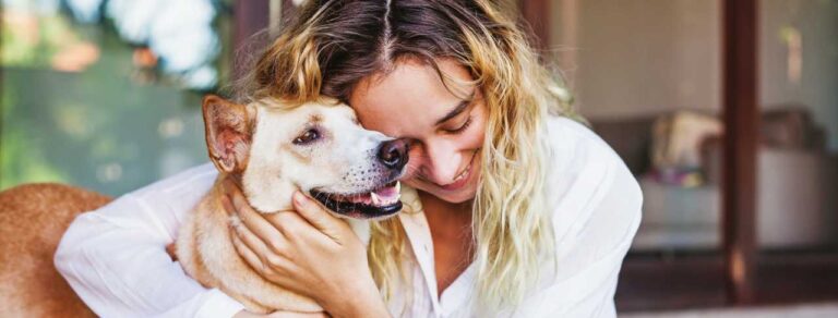 Woman in a white shirt hugs a tan dog, both appear content, indoors with a blurred background.