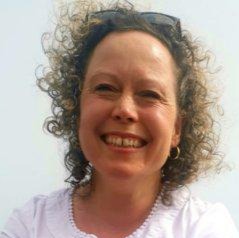 A woman with curly hair and sunglasses on her head is smiling at the camera, wearing a white top and gold hoop earrings, in bright natural light.