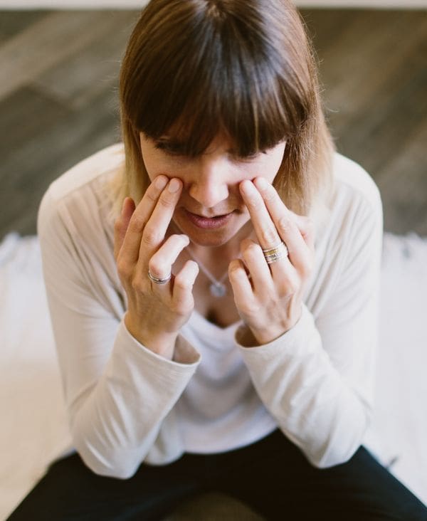 A person sits cross-legged and presses their index and middle fingers to the skin below their eyes, appearing to perform a facial exercise or massage.