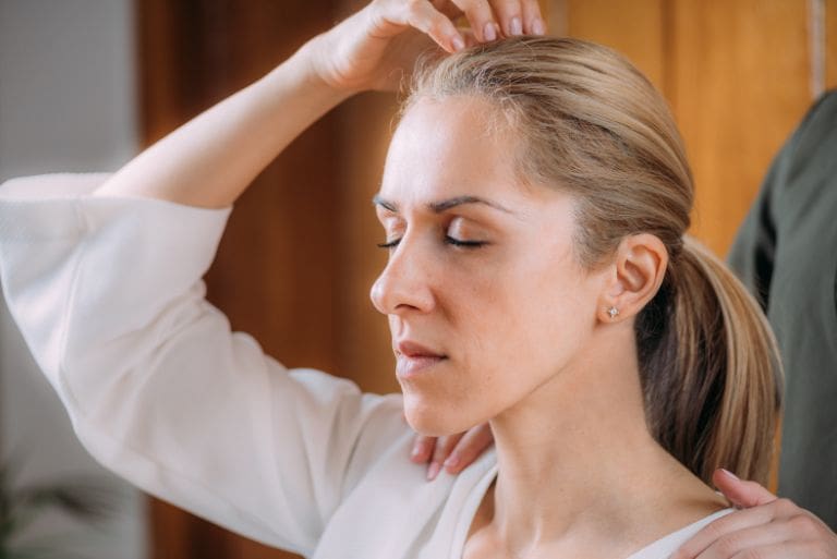A woman with closed eyes has one hand on her head while another person rests a hand on her shoulder in an indoor setting.
