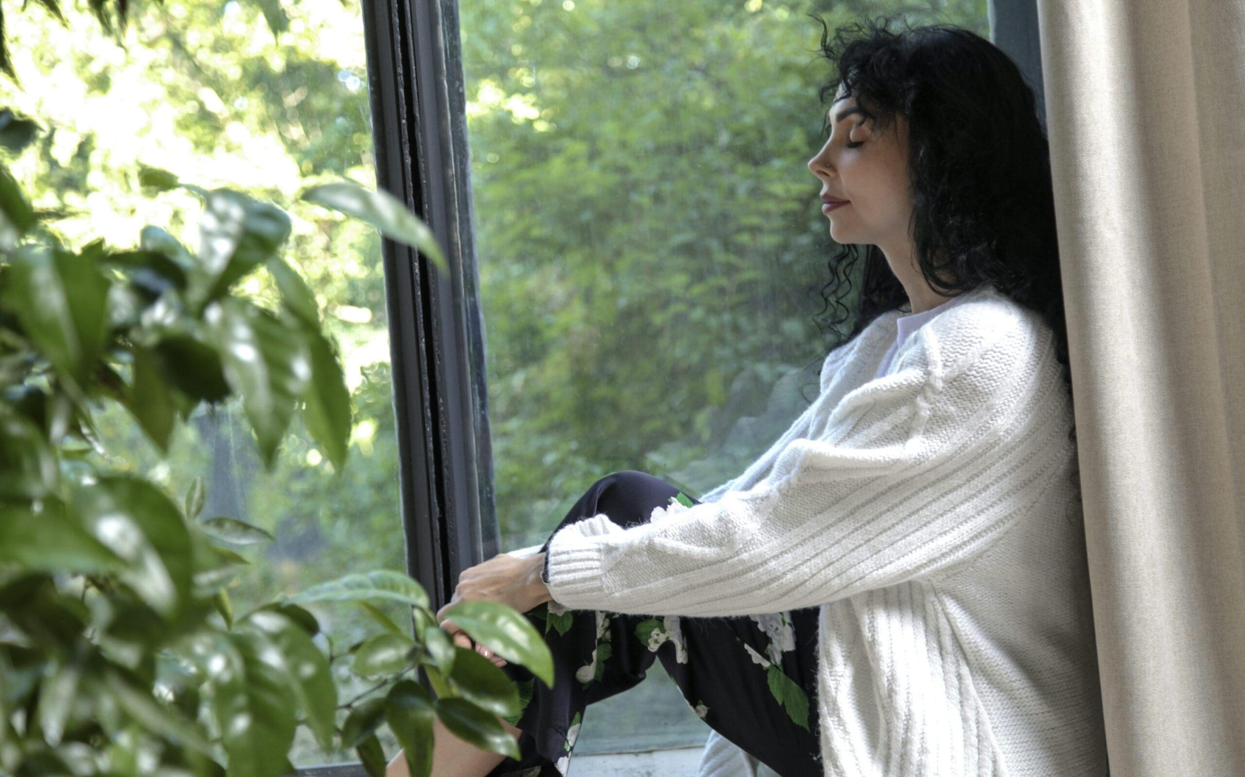 Woman with dark curly hair sits by a window, eyes closed, wearing a white sweater and patterned pants. Green leaves and sunlight are visible outside the window.