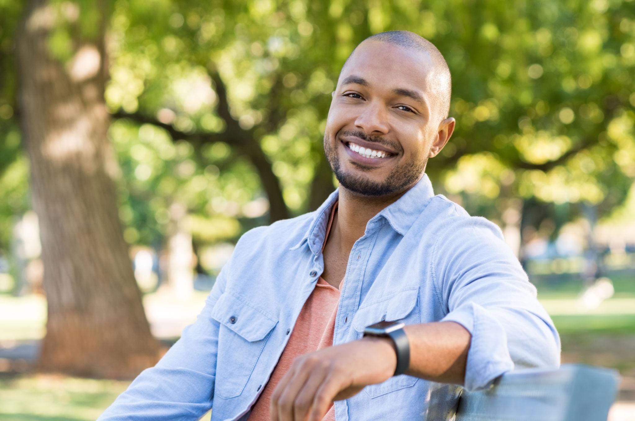 Man with short hair and a beard, wearing a light blue shirt and smartwatch, sits on a bench outdoors, smiling at the camera with trees in the background.