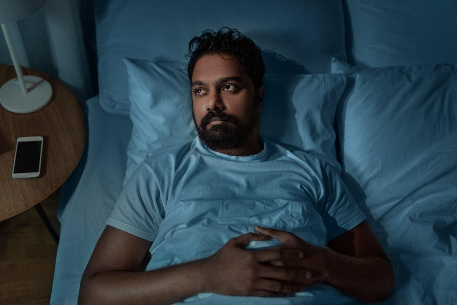 A man lies in bed at night with his hands folded on his chest, looking thoughtful. A smartphone is on the bedside table next to him.