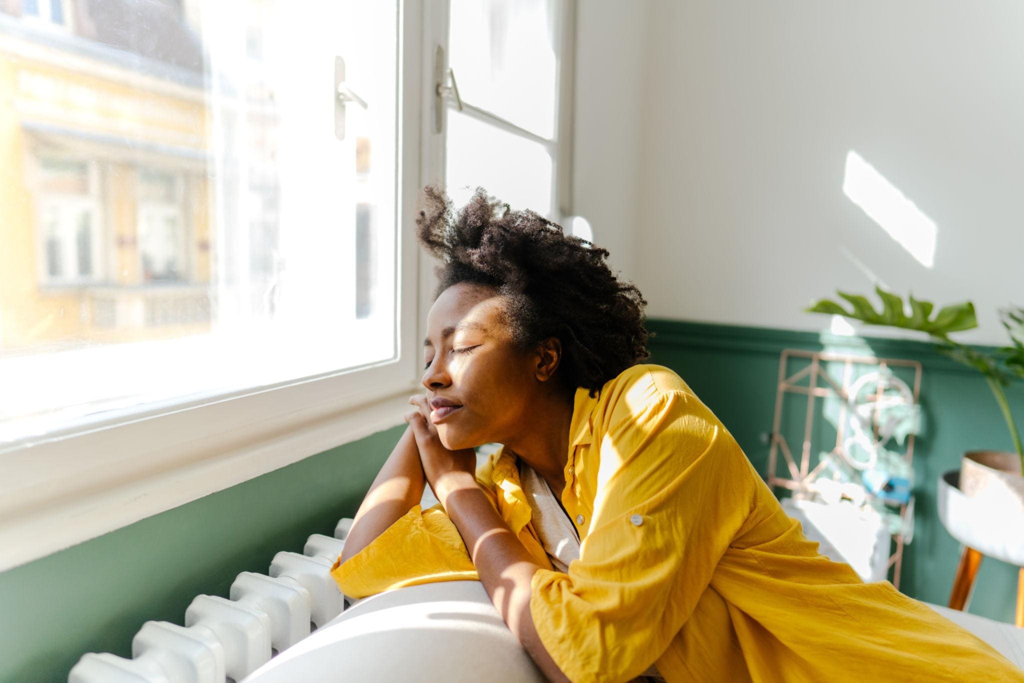 Person in a yellow shirt sits on a couch, resting their head on their hand and looking peaceful while sunlight streams through a nearby window.