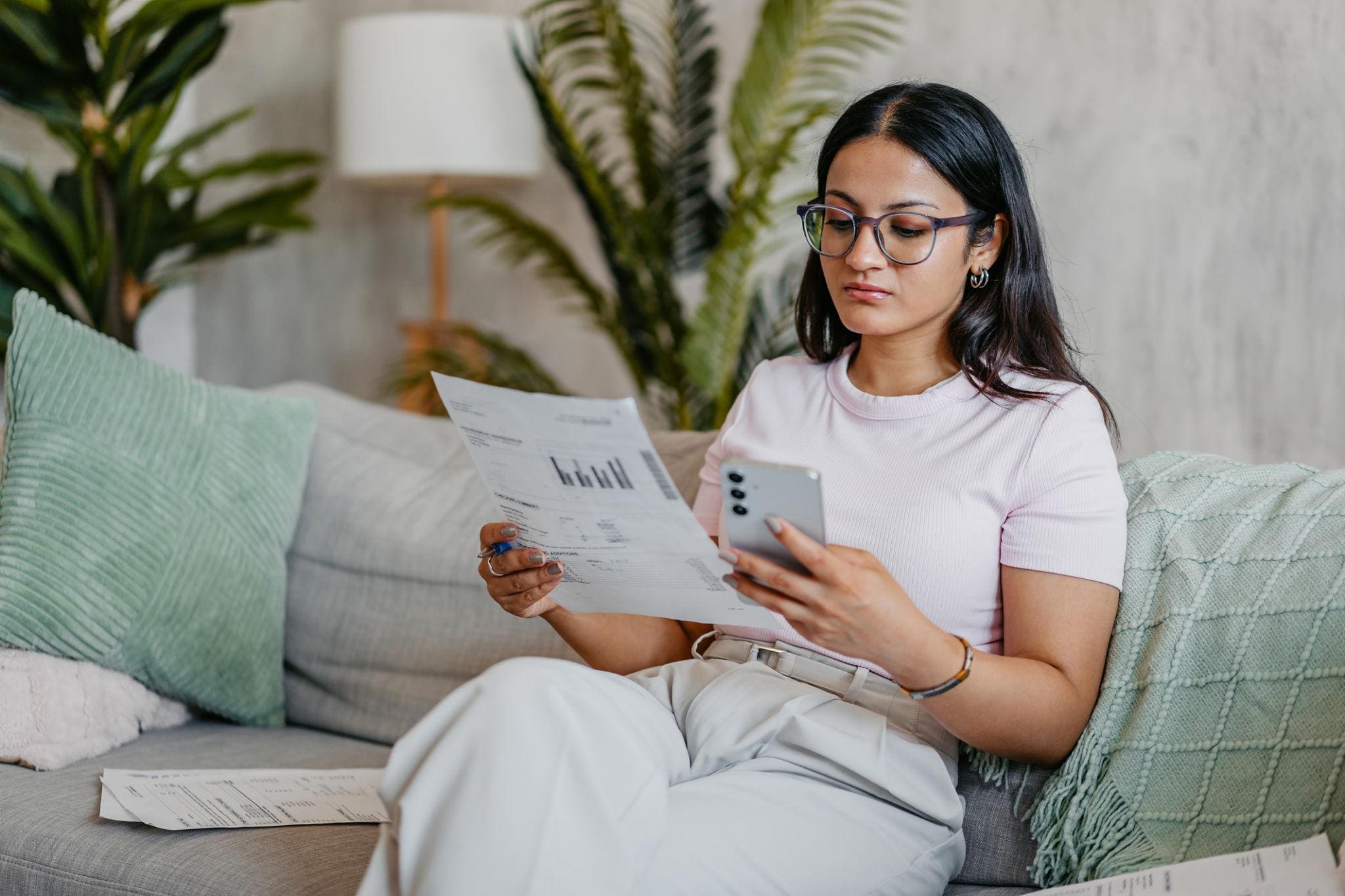 Woman sitting on a couch, holding a piece of paper with graphs in one hand and looking at her smartphone in the other, surrounded by cushions and scattered papers.
